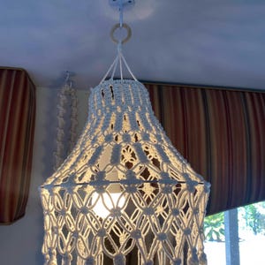 May include: A white macrame lampshade with a light bulb inside. The lampshade is hanging from a white ceiling hook.