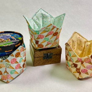 May include: Three fabric ice cream cone-themed containers. One container is holding a pint of Ben & Jerry's ice cream. The containers are made of fabric and have a light blue, yellow, and white background with a pattern of ice cream cones.