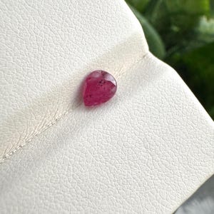 May include: A pear-shaped, faceted gemstone in a deep magenta-pink hue. The stone features a polished surface with visible internal inclusions and a pointed top, resting on a textured, off-white surface with a subtle grain pattern.
