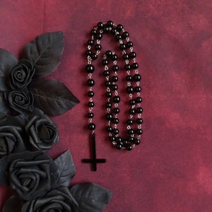 May include: A black beaded necklace with a silver chain and a black upside-down cross pendant. The necklace is on a dark red background.