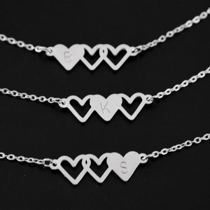 May include: Three silver necklaces with delicate chains and three interlocking heart pendants. Each heart has a different initial engraved inside: B, K, and S.
