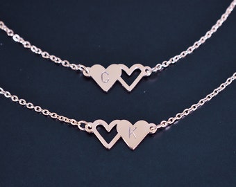 May include: Two rose gold necklaces, each featuring two heart pendants. One necklace has the letter "C" on one heart, the other has the letter "K". The necklaces are displayed on a dark background.