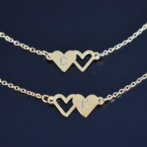 Può includere: Due collane color oro, ciascuna con una catena e due ciondoli a forma di cuore. Una collana ha la lettera "C" su un cuore, e l'altra la lettera "K". Lo sfondo è blu scuro.