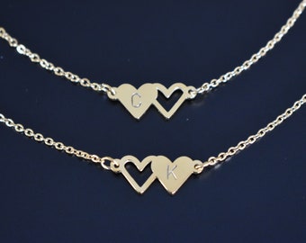 May include: Two gold-colored necklaces, each featuring a chain and two heart pendants. One necklace has the letter "C" on one heart, and the other has the letter "K". The background is a dark blue.
