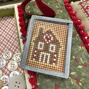 May include: A small, framed cross-stitch ornament featuring a brown house with white details and a red wreath. The ornament has a silver metal frame and a red velvet ribbon for hanging. The background includes fabric with red and green patterns and white buttons.