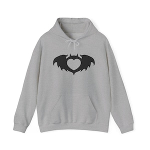 May include: A light grey hooded sweatshirt featuring a black graphic print on the chest. The graphic depicts a heart shape with bat-like wings and small devil horns at the top. The hoodie includes a front kangaroo pocket and matching drawstrings.