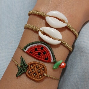 Pode incluir: Quatro pulseiras com diferentes pingentes. As duas primeiras pulseiras têm conchas brancas, a terceira pulseira tem um pingente de melancia vermelho e verde, e a quarta pulseira tem um pingente de abacaxi castanho.