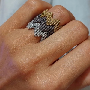 Op de afbeelding: Een zilveren, grijze en gouden gevlochten ring met een zigzagpatroon. De ring wordt gedragen aan een vinger.