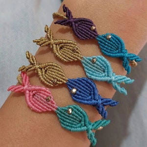 Peut inclure: Cinq bracelets d'amitié en forme de poisson colorés avec un accent perlé sur chaque poisson. Les bracelets sont faits de ficelle nouée et sont superposés au poignet d'une personne.