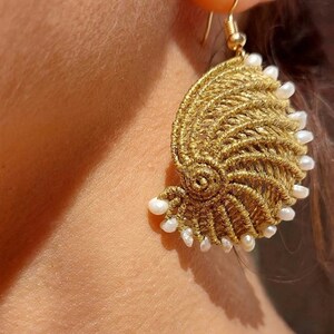 May include: A gold-colored macrame earring in a spiral seashell design, adorned with small white pearls along the edge. The earring is suspended from a gold-tone hook. The jewelry is displayed close-up against a blurred background.