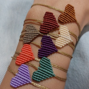 Puede incluir: Un conjunto de ocho pulseras de la amistad con dijes en forma de corazón en varios colores, incluyendo morado, verde, naranja, rojo, marrón, beige y crema. Las pulseras están hechas con un hilo de color dorado.