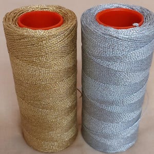 Peut inclure: Deux bobines de fil métallique, une dorée et une argentée, chacune avec un noyau en plastique rouge. Le fil est étroitement enroulé, formant une forme cylindrique. L'image en gros plan met en évidence la texture et l'éclat du fil.