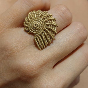 Op de afbeelding: Een gouden ring met een spiraalvormig ontwerp, dat doet denken aan een schelp. De ring is gemaakt van geweven draad en heeft een uniek, ingewikkeld patroon.