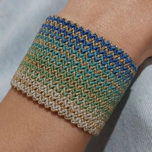 Peut inclure: Un bracelet tissé avec un dégradé de couleurs bleu, vert et beige. Le bracelet est tissé avec un fil doré.