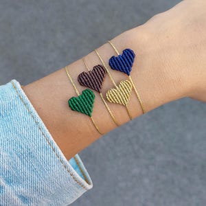 Op de afbeelding: Vier macramé hart armbanden in groen, bruin, marineblauw en goud. Elk hart is bevestigd aan een delicate gouden ketting. De armbanden worden gedragen om een pols tegen een grijze achtergrond.