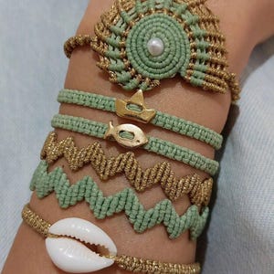 Può includere: Un set di cinque braccialetti con uno schema di colori verde e oro. I braccialetti sono realizzati con corda intrecciata e presentano vari ciondoli, tra cui una conchiglia, una corona e un pesce.