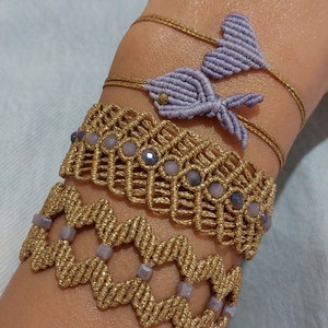 Puede incluir: Un conjunto de cuatro pulseras con una combinación de colores dorado y morado. Las pulseras están hechas de cordón trenzado y presentan varios patrones y cuentas. Dos de las pulseras tienen un diseño de pez.