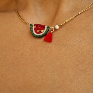 Pode incluir: Um colar com um pequeno pingente de melancia semicircular feito de fio vermelho, branco e verde. O pingente inclui pequenos detalhes quadrados em tom dourado, uma pequena conta branca e uma borla vermelha pendurada num fio fino dourado.