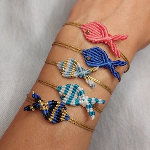 Pode incluir: Cinco pulseiras de amizade coloridas com pingentes de peixe. As pulseiras são feitas com fio dourado e apresentam pingentes de peixe de diferentes cores, incluindo azul, rosa, branco e preto.