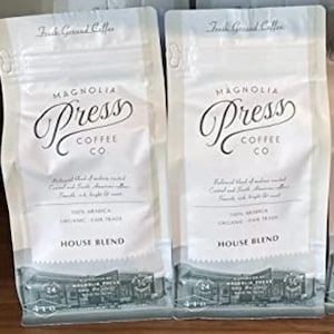 May include: Two white coffee bags with the text "Fresh Ground Coffee" at the top. The bags feature the "Magnolia Press Coffee Co." logo in elegant script. The bags are labeled "House Blend" and contain 100% Arabica coffee.