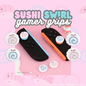 May include: Two black game controllers with blue and orange side rails, featuring white thumbstick covers shaped like sushi swirls with pink or blue centers and small smiling faces. The text SUSHI SWIRL gamer grips is displayed above.