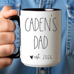 May include: White ceramic mug with a black interior and handle. The mug features the text "CADEN'S DAD" in a black, handwritten-style font, with "est. 2026" below. A small black heart is next to the date.