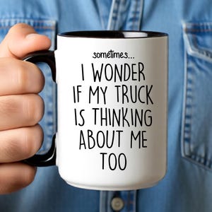 May include: A white ceramic mug with a black interior and handle. The mug features the text "sometimes... I WONDER IF MY TRUCK IS THINKING ABOUT ME TOO" in black lettering. The mug is held by a person wearing a blue denim shirt.
