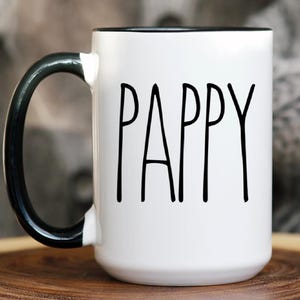 May include: White ceramic mug with a black handle and rim. The word "PAPPY" is printed in black, minimalist font on the front of the mug. The mug is sitting on a wooden surface.