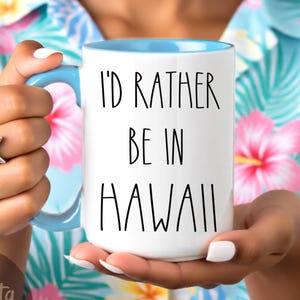 May include: White ceramic mug with a blue interior and handle, featuring the text "I'D RATHER BE IN HAWAII" in black. The mug is held against a floral background, with a starfish ring on the hand.