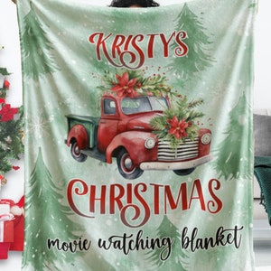 May include: A light green Christmas-themed blanket with a red vintage truck illustration. The truck is decorated with a Christmas tree and flowers. Text on the blanket reads "Kristy's Christmas movie watching blanket."