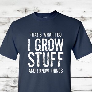 May include: Navy blue t-shirt with white text that reads "THAT'S WHAT I DO I GROW STUFF AND I KNOW THINGS." The shirt is a short-sleeved crew neck style.