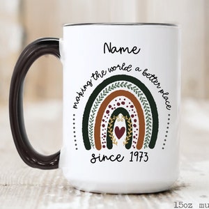 May include: White 15oz mug with a black handle and rim. The mug features a rainbow design with the text "making the world a better place" and "since 1973". The word "Name" is above the rainbow.