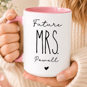 Puede incluir: Una taza de cer&aacute;mica blanca con interior y asa de color rosa suave. El frente presenta el texto negro Future MRS. Powell con un peque&ntilde;o icono de coraz&oacute;n negro centrado debajo del nombre. El dise&ntilde;o est&aacute; impreso en una fuente simple y limpia.