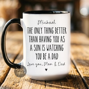 May include: A white ceramic mug with a black handle and rim, featuring the black printed text: Michael, THE ONLY THING BETTER THAN HAVING YOU AS A SON IS WATCHING YOU BE A DAD, Love you, Mom & Dad, with a small heart icon below the text.