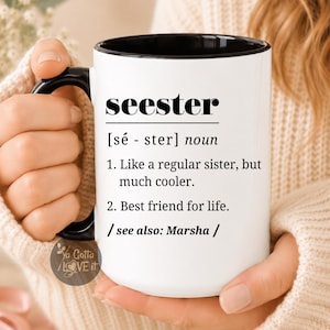 May include: A white ceramic mug with a black interior and handle, featuring black printed text that defines the word seester as a noun, describing it as a cooler version of a sister and a best friend for life, with a see also Marsha reference.