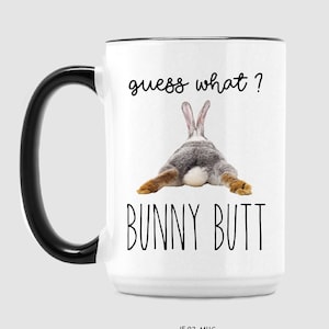 Puede incluir: Taza de cer&aacute;mica blanca con asa y borde negros. La taza presenta el texto "guess what?" encima de una imagen del trasero de un conejo, con las palabras "BUNNY BUTT" debajo. La taza est&aacute; etiquetada como "15OZ MUG".