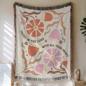 May include: Woven throw blanket with a floral design in shades of orange, pink, and brown. The blanket features the text "TRUST IN THE LORD WITH ALL YOUR HEART" in a brown script. The edges are finished with a fringed trim.