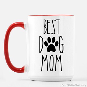 May include: White ceramic mug with a red rim and handle. The mug has black text that reads "BEST DOG MOM" with a paw print in the middle of the word "DOG".
