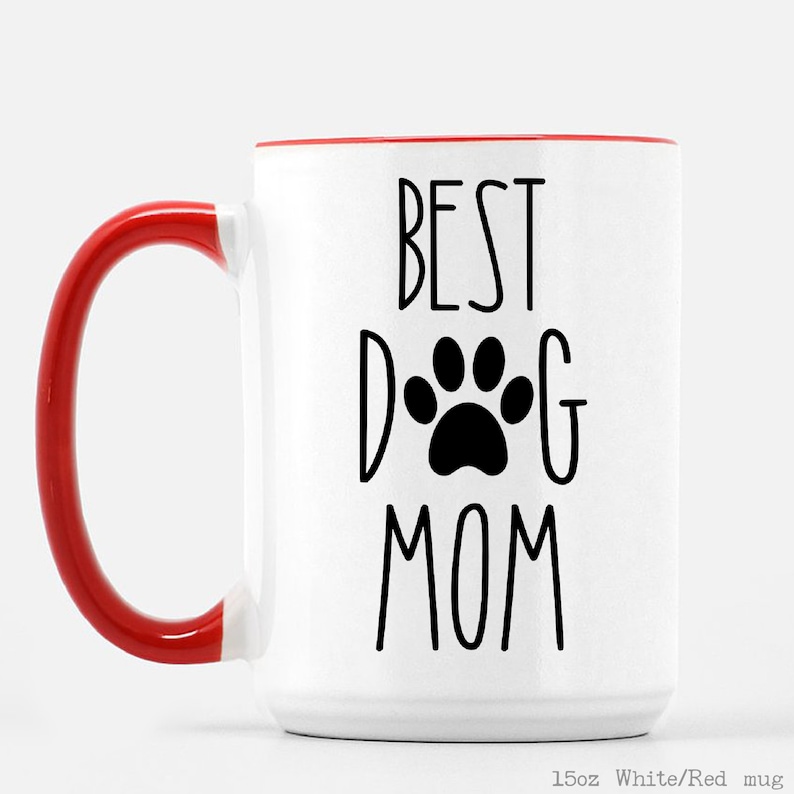May include: White ceramic mug with a red rim and handle. The mug has black text that reads "BEST DOG MOM" with a paw print in the middle of the word "DOG".