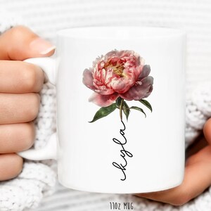 May include: White ceramic mug with a pink peony flower and the name "Kayla" written in cursive on the front. The mug is 11 oz.