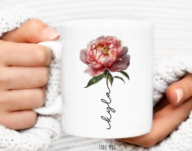 May include: White ceramic mug with a pink peony flower and the name "Kayla" written in cursive on the front. The mug is 11 oz.