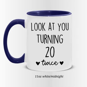 May include: White ceramic mug with a navy blue handle and rim. The mug features the text "LOOK AT YOU TURNING 20 twice" in black, with two black heart symbols. The mug is 11oz.