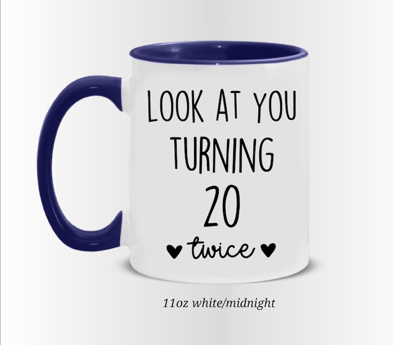 May include: White ceramic mug with a navy blue handle and rim. The mug features the text "LOOK AT YOU TURNING 20 twice" in black, with two black heart symbols. The mug is 11oz.