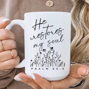 May include: A white ceramic mug featuring black text that reads He restores my soul and Psalm 23:3. Below the text is a black line art illustration of various wildflowers and stems. The mug has a smooth finish and a curved handle on the left side.