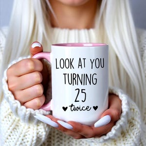 May include: White ceramic mug with a pink interior and handle. The mug features the text "LOOK AT YOU TURNING 25 twice" in black, with two black heart symbols. The mug is held by a person wearing a white sweater.