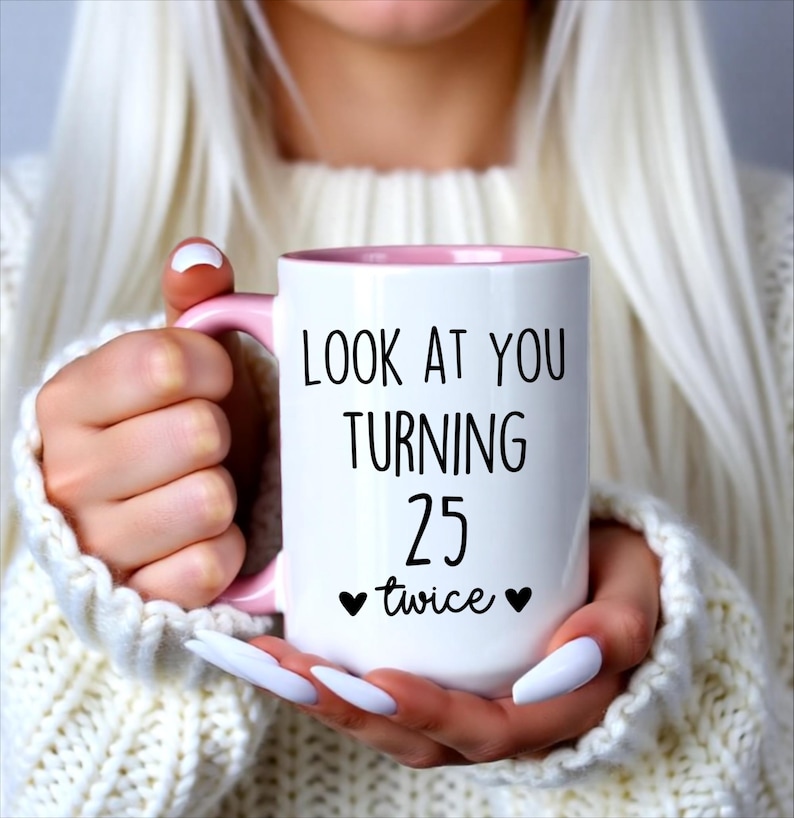 May include: White ceramic mug with a pink interior and handle. The mug features the text "LOOK AT YOU TURNING 25 twice" in black, with two black heart symbols. The mug is held by a person wearing a white sweater.