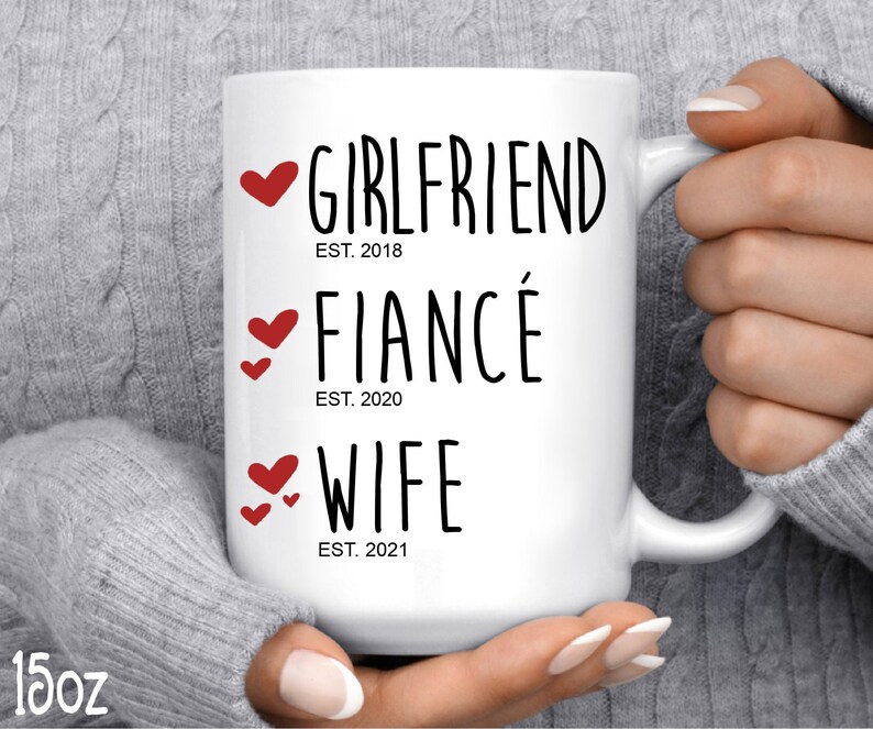 以下が含まれることがあります： 白いセラミック製のマグカップに、黒い文字で「Girlfriend Est. 2018、Fianc&eacute; Est. 2020、Wife Est. 2021」と書いてあります。文字は小さな赤いハートで飾られています。