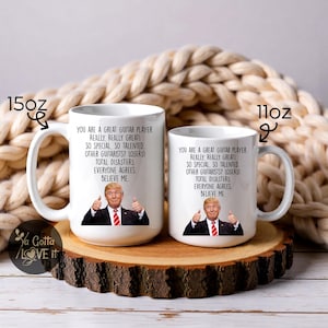 Puede incluir: Dos tazas de cer&aacute;mica blanca con texto negro y la imagen de un hombre haciendo una se&ntilde;al de pulgar hacia arriba. El texto dice: "ERES UN GRAN GUITARRISTA. &iexcl;REALMENTE, REALMENTE GENIAL!" Una taza es de 425 ml, la otra de 312 ml. Las tazas est&aacute;n sobre una superficie de madera.