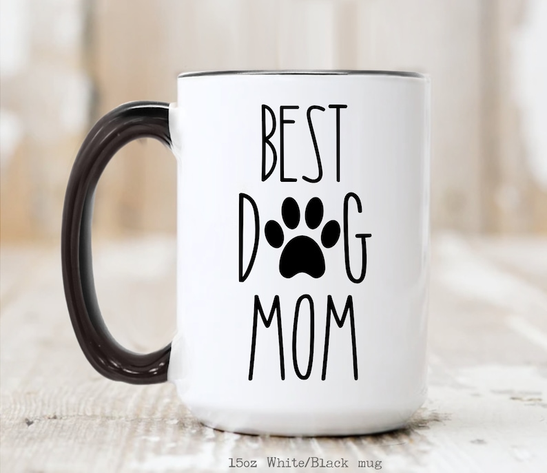 May include: White ceramic mug with a black handle and black rim. The mug has the text "BEST DOG MOM" in black lettering with a black paw print in the center of the "D" and "G".