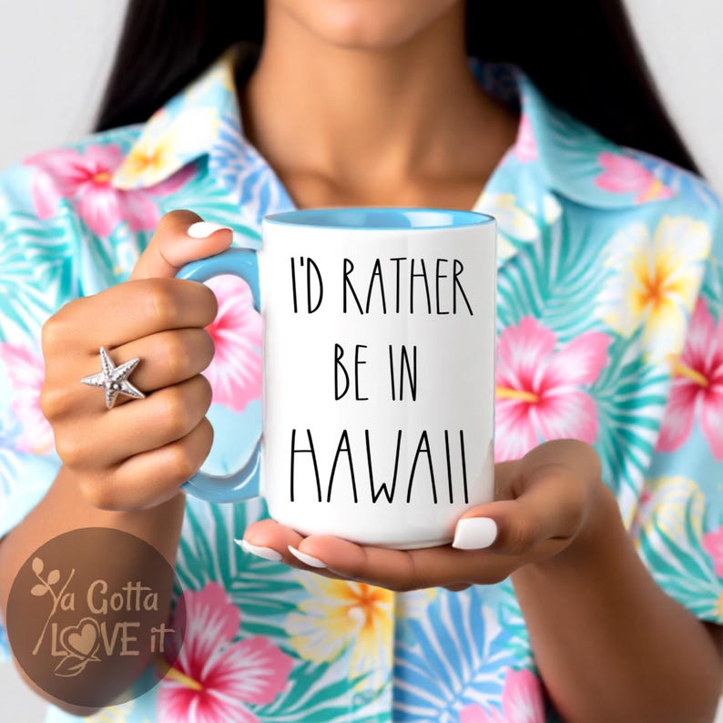 May include: White ceramic mug with a blue interior and handle, featuring the text "I'D RATHER BE IN HAWAII" in black. The mug is held against a floral background, with a starfish ring on the hand.